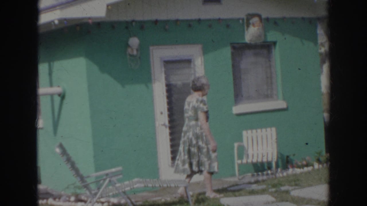 The woman walks toward the green house exterior, with a slatted chair, door, window, and lawn furniture visible.