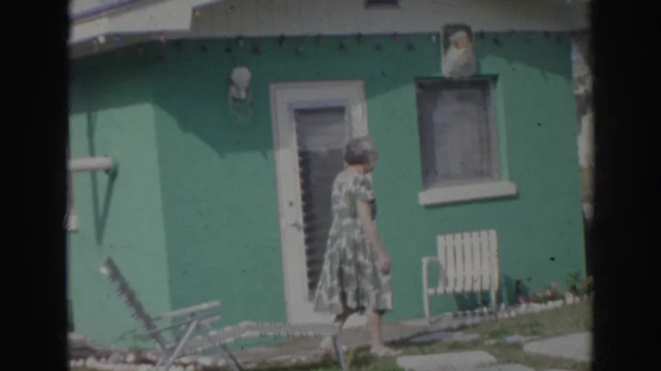 The woman walks toward the green house exterior, with a slatted chair, door, window, and lawn furniture visible.