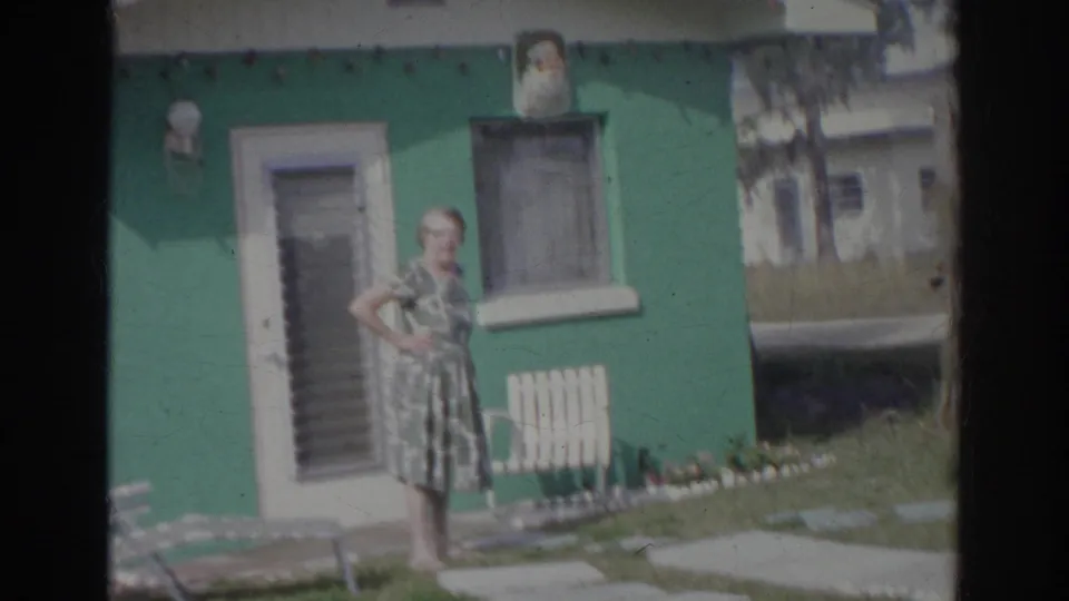 In the final frame, the woman stands with a hand on her hip near the green Norfolk house and yard furniture.