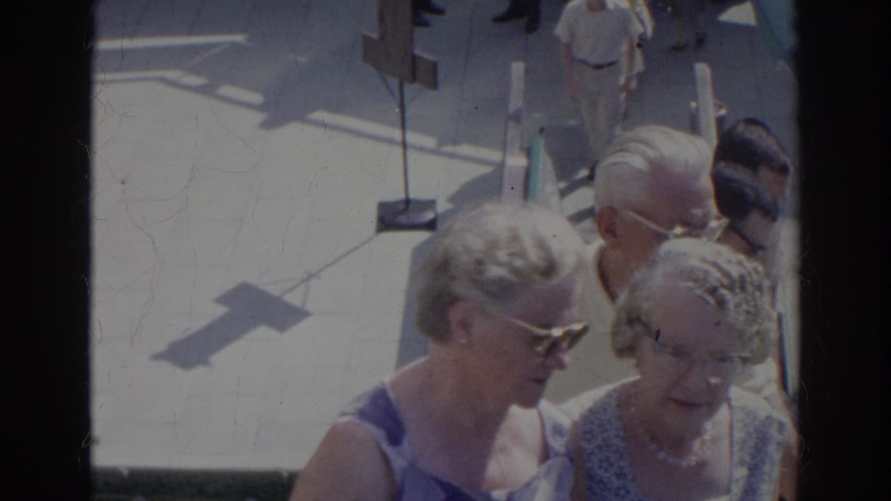 A small group of older women walks outdoors in the 1966 Norfolk footage, seen from above with bright sun and deep frame-edge shadows.