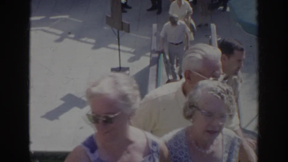 1966 Norfolk home-movie frame showing older adults on sunlit outdoor stairs, including two women in sunglasses near the foreground.