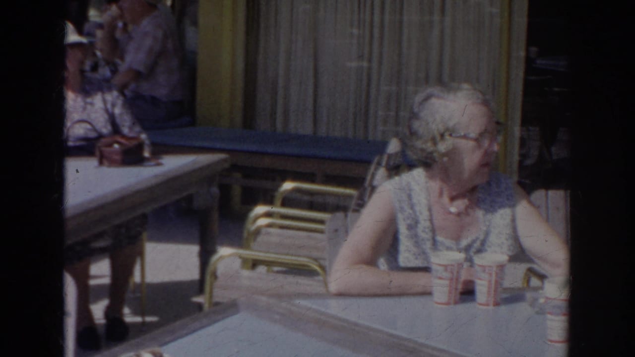 Elderly woman seated at an outdoor cafe table with paper cups nearby.