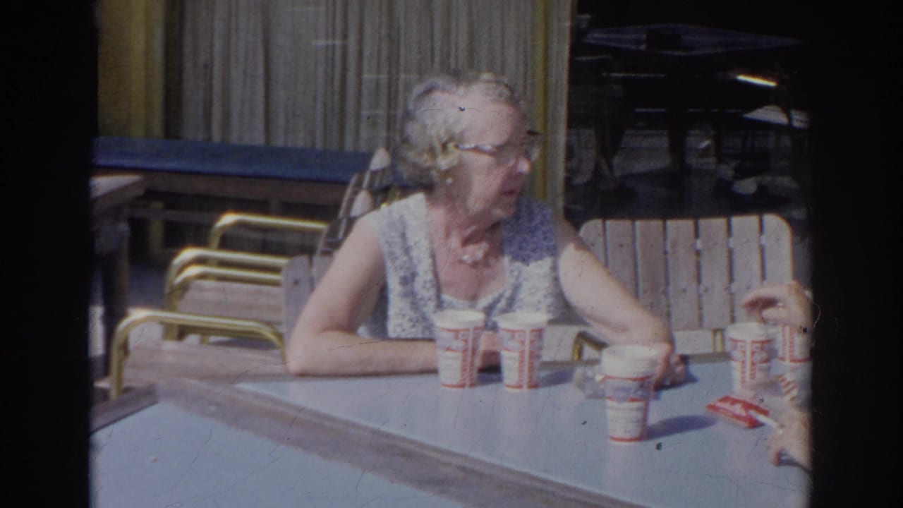An elderly woman sits at an outdoor cafe table in 1966 Norfolk, with several paper cups and snack packaging in front of her.