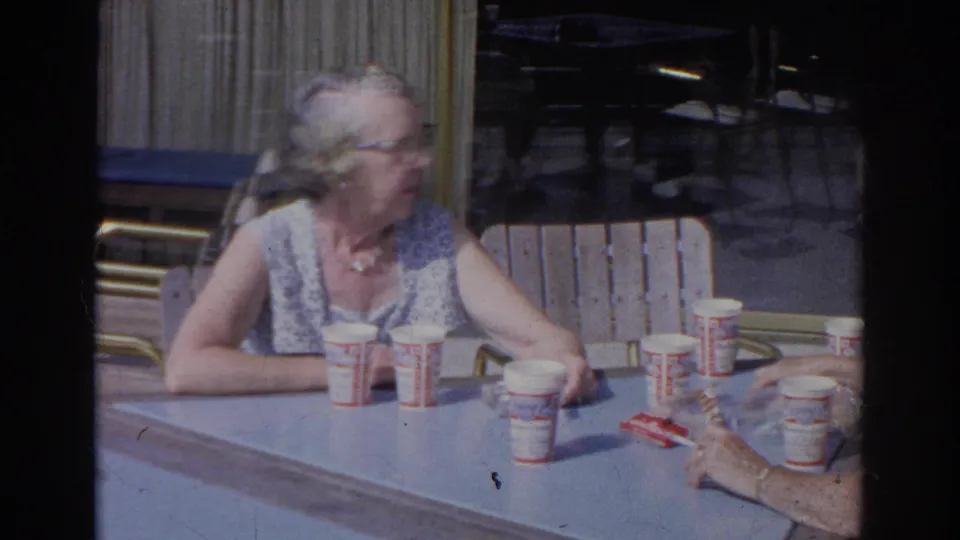 At a Norfolk outdoor cafe, an elderly woman looks across the table as another person's hand reaches among paper cups and snacks.