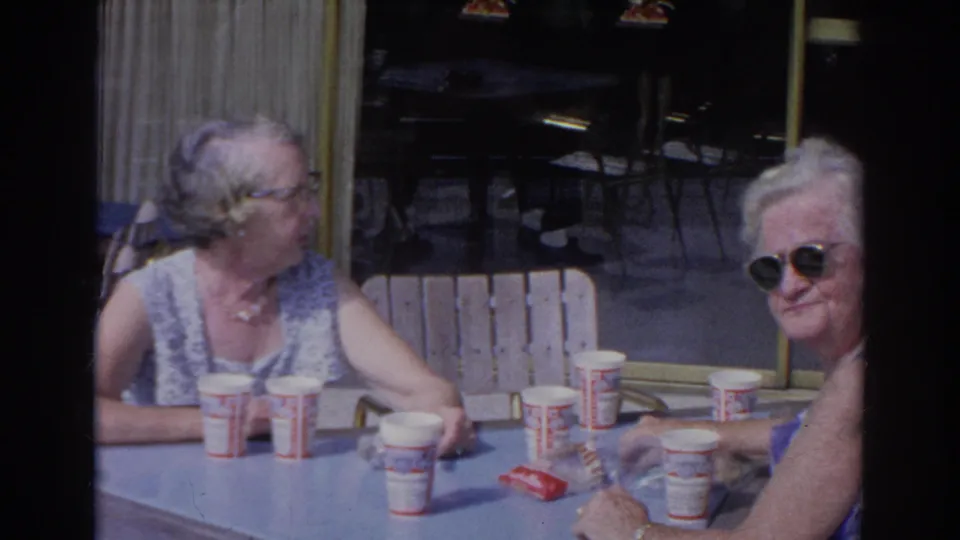 Two elderly women sit at a Norfolk cafe table in 1966, with cups and snack items on the blue tabletop.