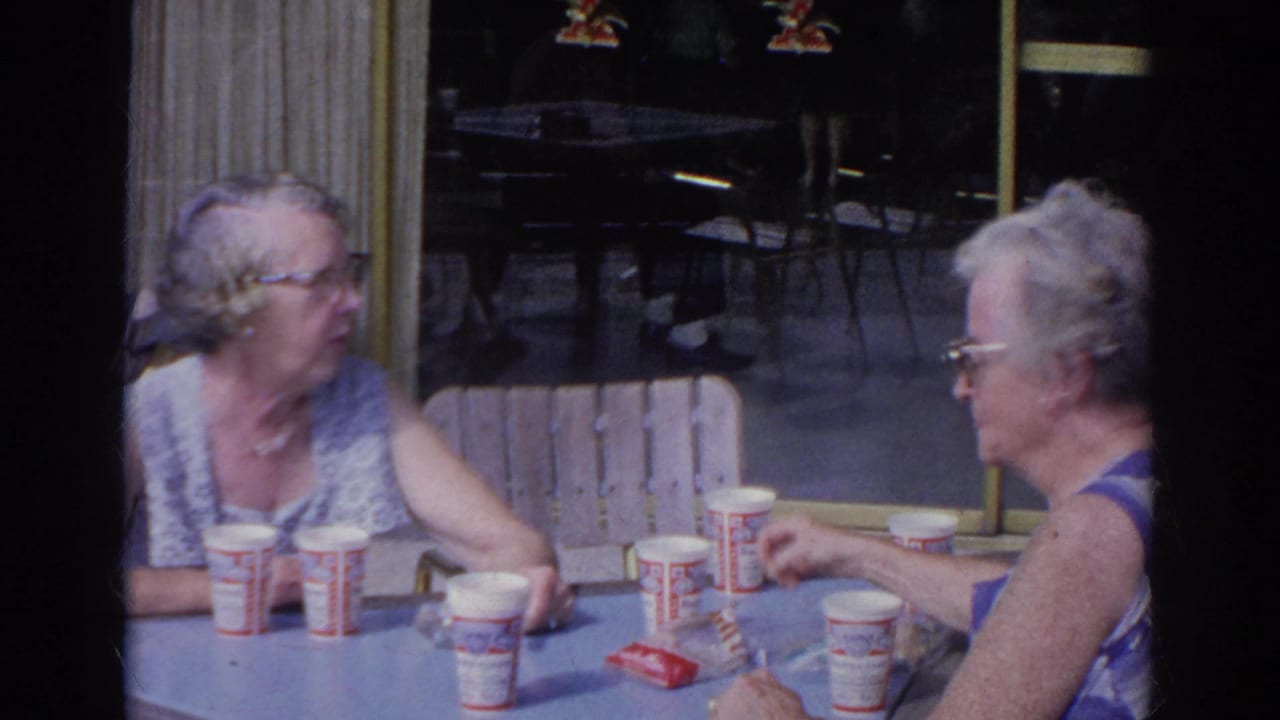 Two older women sit at a diner table with paper cups and food items.