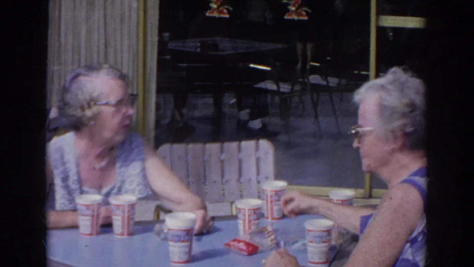 In 1966 Norfolk home-movie footage, two older women sit at an outdoor diner table with paper cups and a red packet.