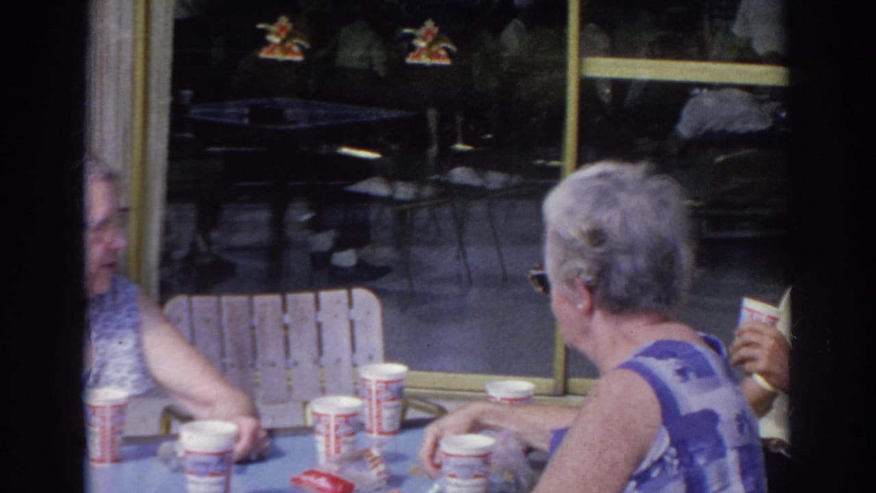 Two older women converse at a diner table in Norfolk, Virginia, with branded paper cups and reflective windows behind them.