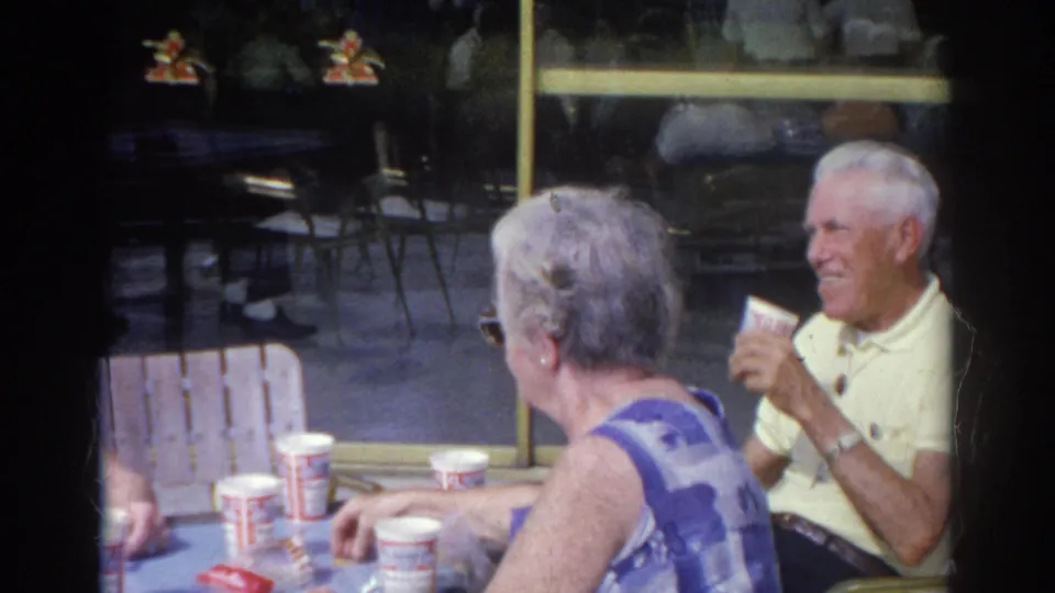 An older woman in sunglasses sits near the camera as an older man in a yellow polo shirt drinks from a paper cup.