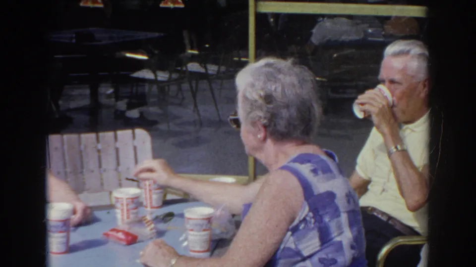 The older man drinks from a paper cup while an older woman in a blue patterned sleeveless top sits beside the diner table.
