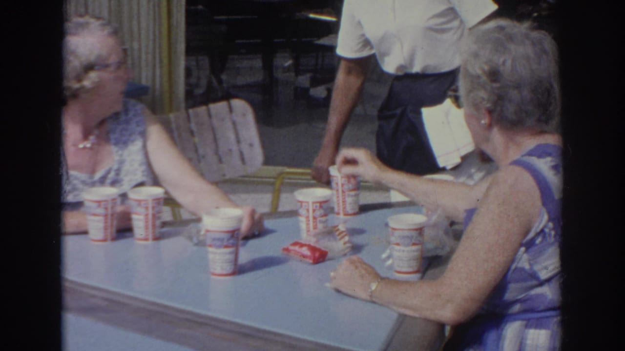 1966 Norfolk diner scene with older patrons at a blue table, a server in white and dark apron, and branded paper cups visible.