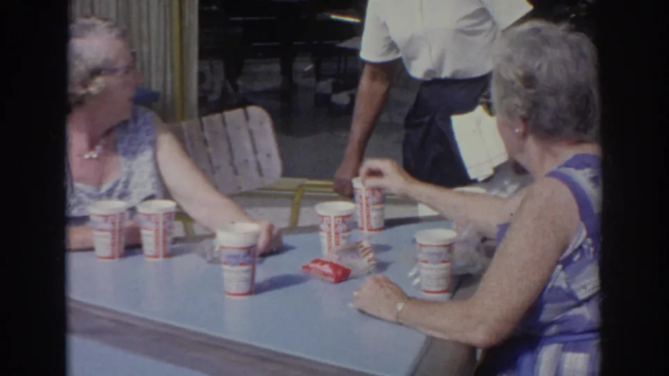 1966 Norfolk diner scene with older patrons at a blue table, a server in white and dark apron, and branded paper cups visible.