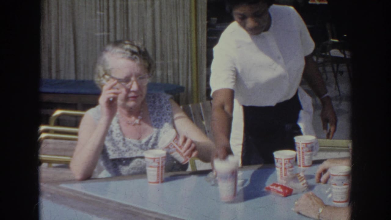1966 Norfolk home-movie frame of an older woman adjusting glasses at a diner table while a server passes beside her.