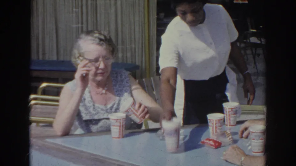 1966 Norfolk home-movie frame of an older woman adjusting glasses at a diner table while a server passes beside her.