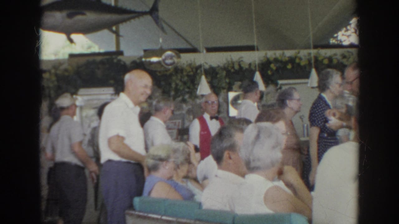 1966 Norfolk social mixer with older adults moving through a decorated interior and a waiter in a red vest visible among the crowd.