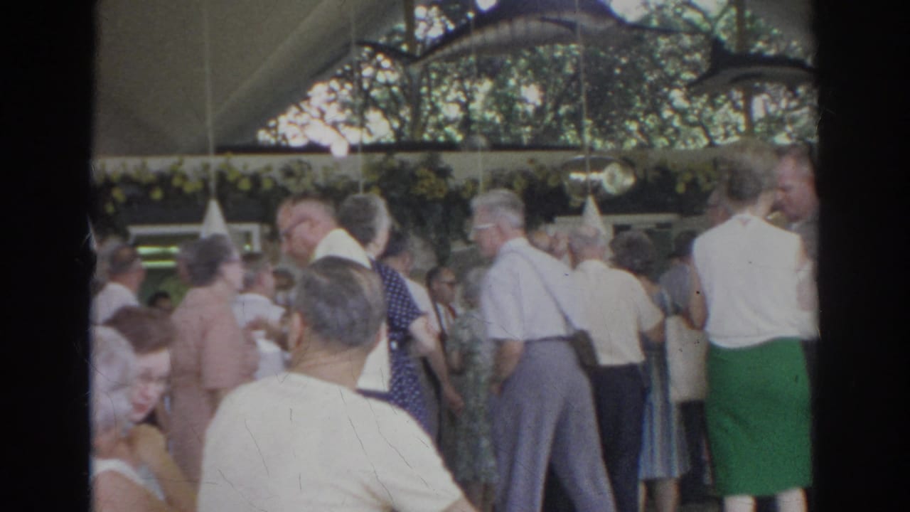 1966 Norfolk home-movie frame showing adults in 1960s clothing mingling with cups beneath a decorated pavilion canopy.