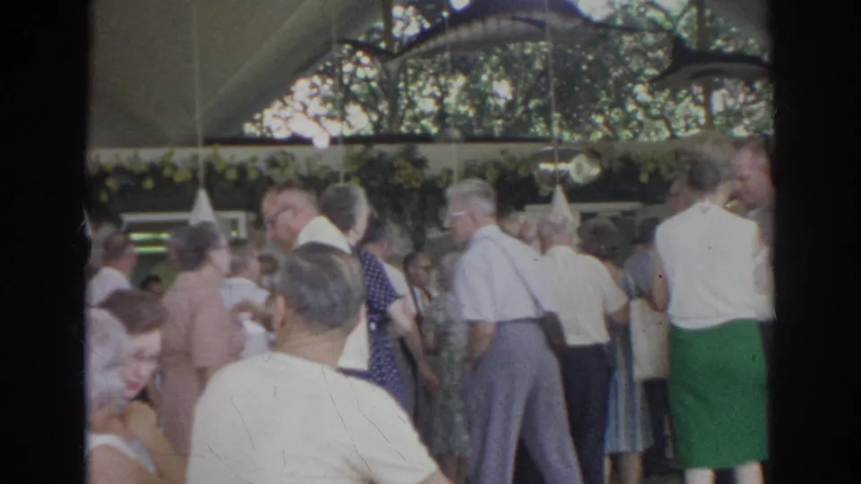1966 Norfolk home-movie frame showing adults in 1960s clothing mingling with cups beneath a decorated pavilion canopy.