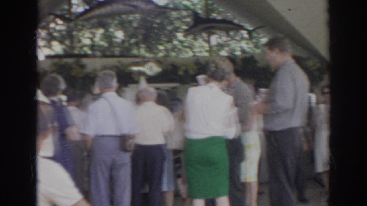 Crowd of adults under a canopy, with a man at right holding a cup.