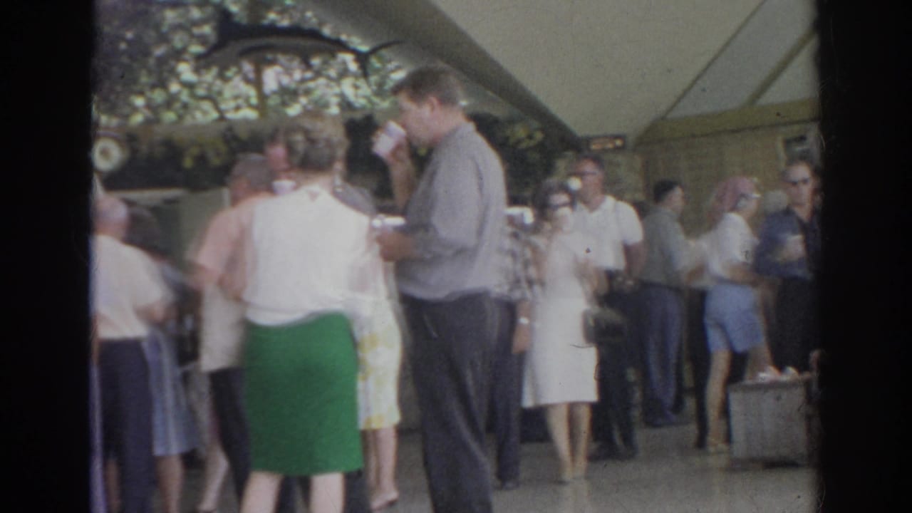 Norfolk 1966 home-movie frame of adults socializing inside a pavilion, including women in dresses and a man drinking from a cup.
