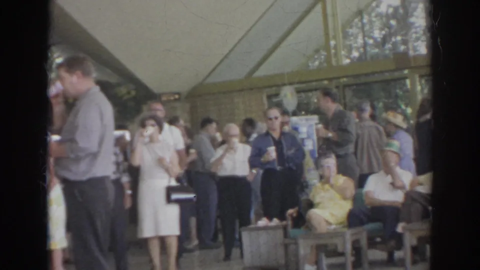 1966 Norfolk social scene with adults seated and standing under a pavilion canopy, several holding drinks in a candid home-movie view.
