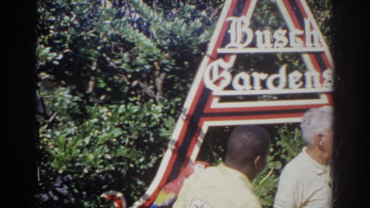 In 1966 Norfolk, Virginia, a man in a white shirt and a yellow-uniformed employee stand near macaws before a Busch Gardens sign.