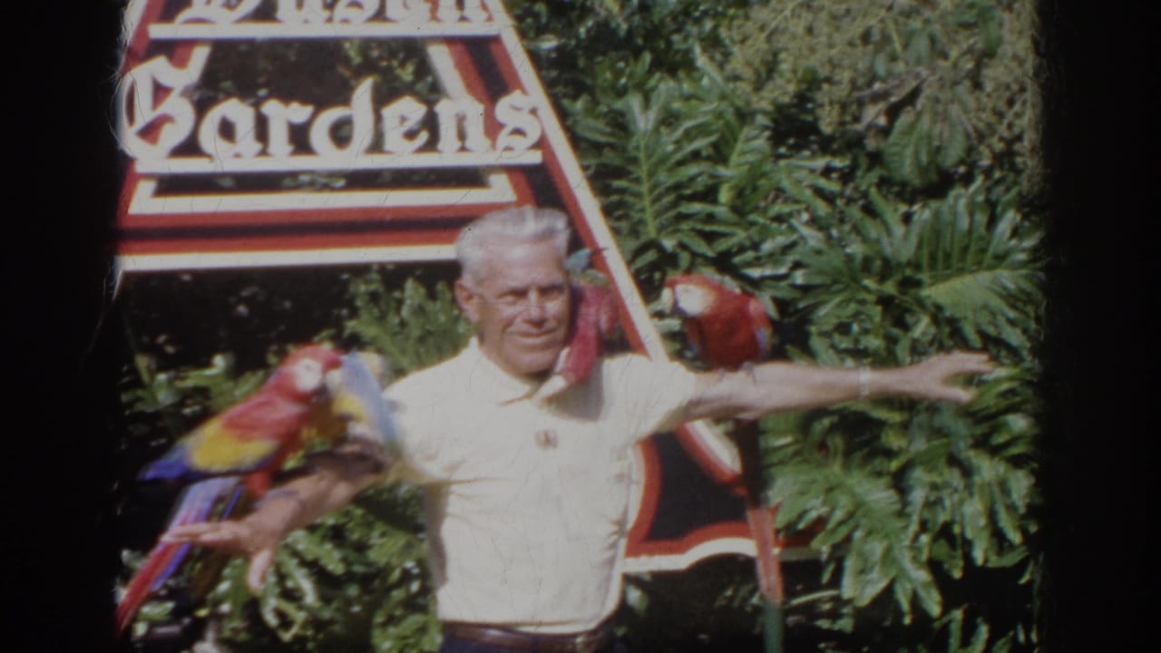 1966 Norfolk home-movie frame of a man with colorful macaws perched on his arms near a Busch Gardens sign.