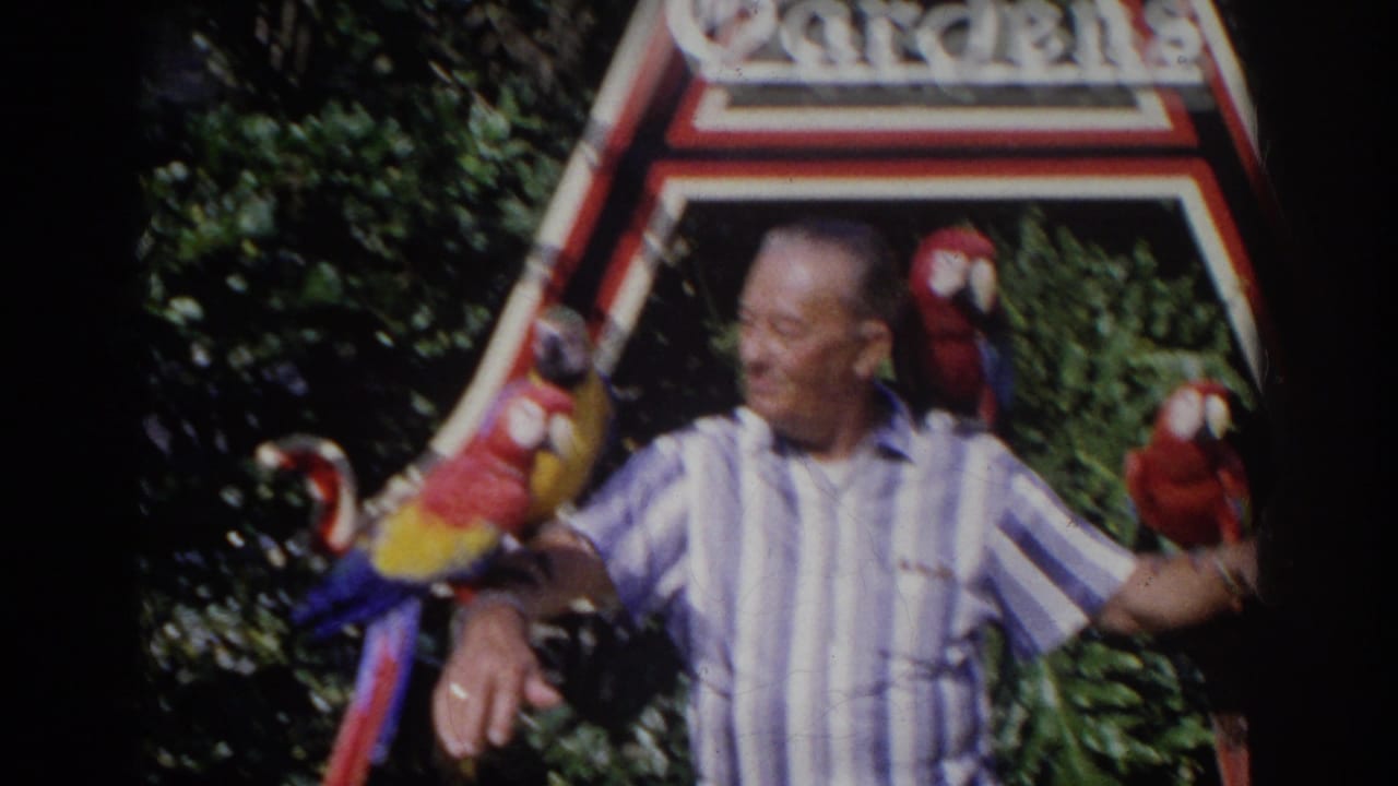 Man in striped shirt holding macaws near the park sign.