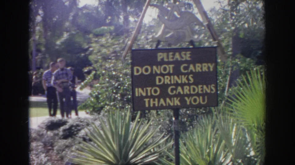 A posted zoo garden sign reads not to carry drinks into the gardens, with visitors and dense planting nearby.