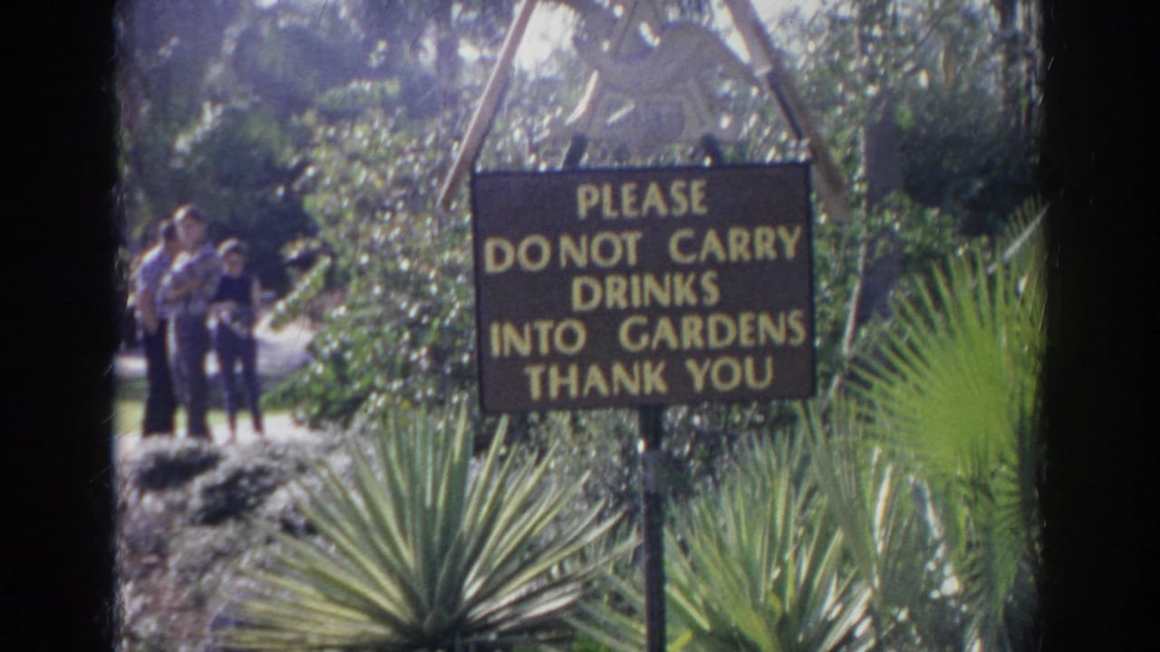 Brown sign with yellow lettering beside garden plants and people on a path.