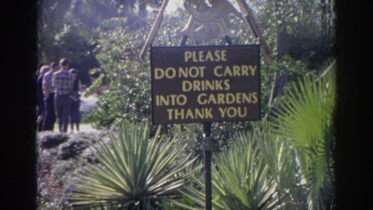 Brown and yellow garden rules sign in Norfolk, Virginia, with adults walking on a path among landscaped plants.