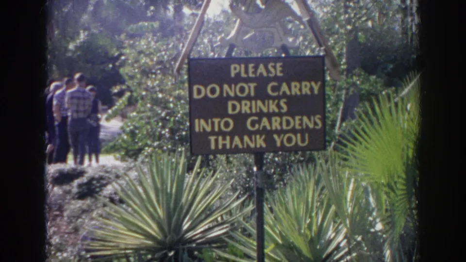 Brown and yellow garden rules sign in Norfolk, Virginia, with adults walking on a path among landscaped plants.