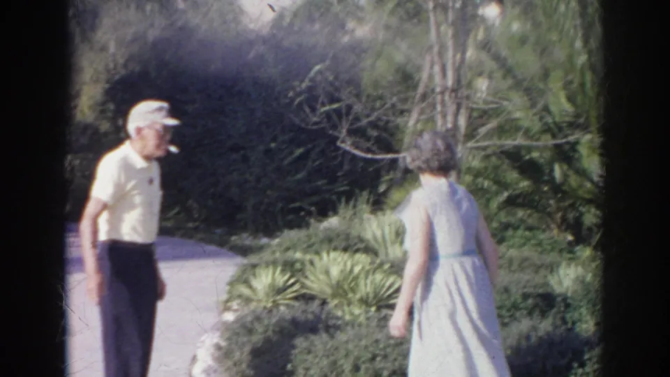 Adults in casual 1960s clothing walk through a Norfolk garden path bordered by shrubs and spiky plants.