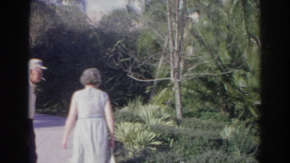 Norfolk garden path scene with a woman in a light dress walking away through lush plantings and a man at the edge of frame.
