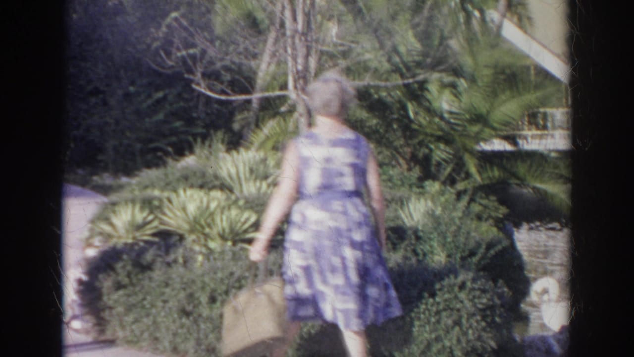 A woman in a purple patterned dress carries a handbag along a garden path in the Norfolk, Virginia home-movie scene.