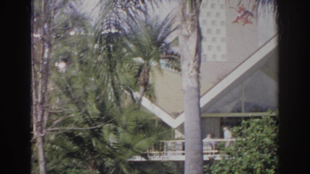 Palm trees partly obscure a pale building with a sharp triangular roofline and porch railing.