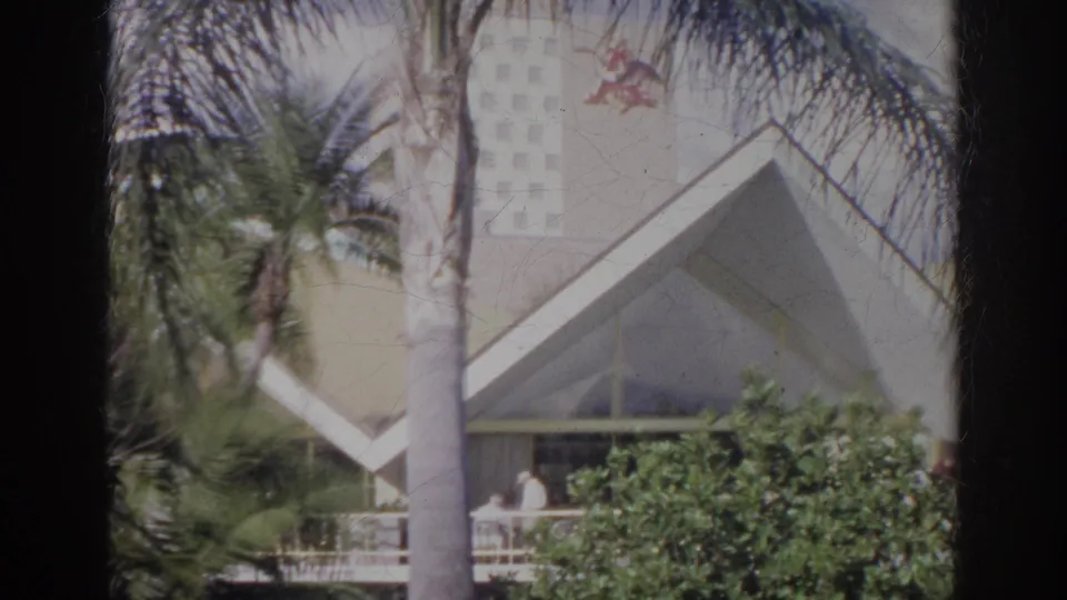 1966 Norfolk exterior view with palms, angular architecture, greenery, and a distant person on the porch.