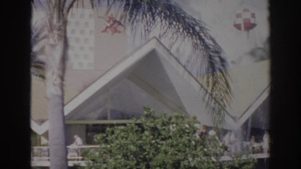 1966 Norfolk 8mm frame showing a sunlit angular exterior, palm foliage, shrubs, and a partially visible porch.