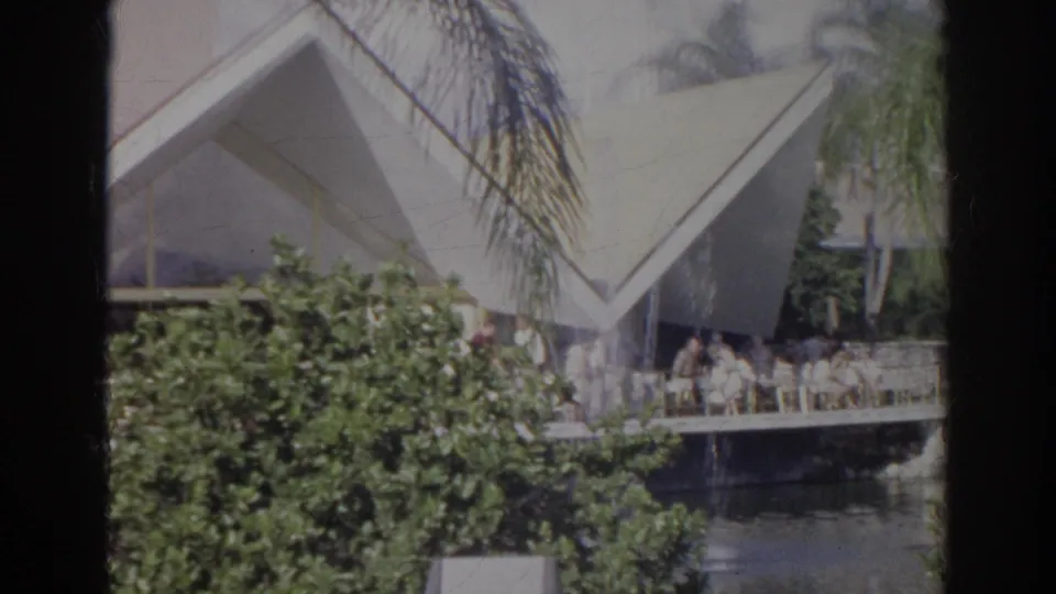 1966 Norfolk home-movie frame with distant porch visitors, dense greenery, palms, and modern angular exterior architecture.
