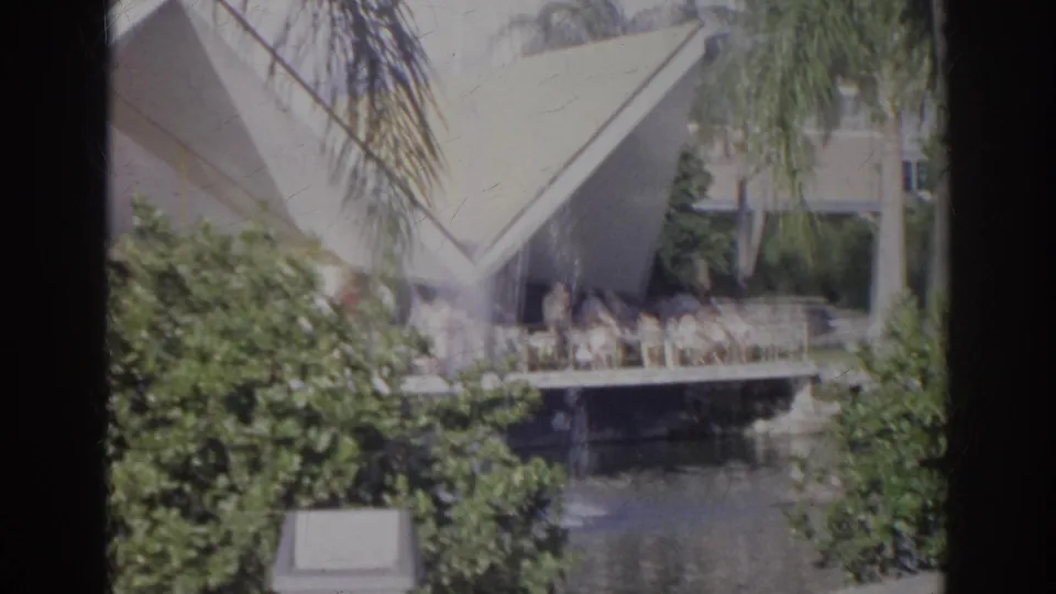 1966 Norfolk home-movie frame showing a landscaped pond, waterfowl, dense shrubs, palms, and an angular modernist building beyond the water.
