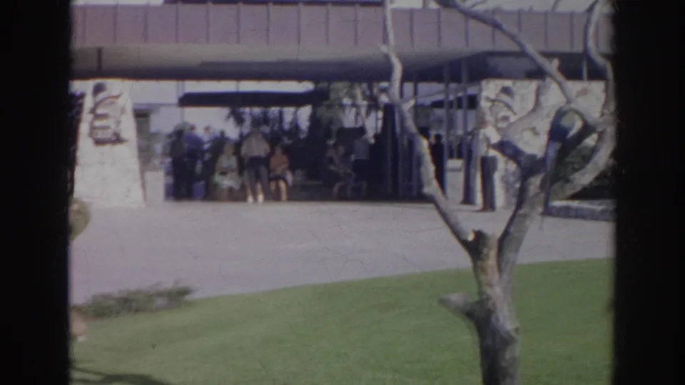 1966 Norfolk home-movie view of a lawn and tree before a modern entrance where several people stand under a covered walkway.