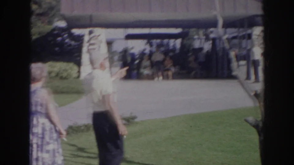 1966 Norfolk 8mm frame showing an older couple crossing a grassy yard before a modern building with people near the entrance.