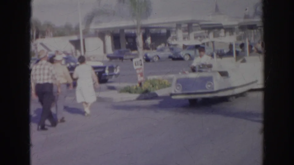 1966 Norfolk home-movie frame with visitors, classic cars, a roadside building, and a small boat-shaped ride at an attraction.