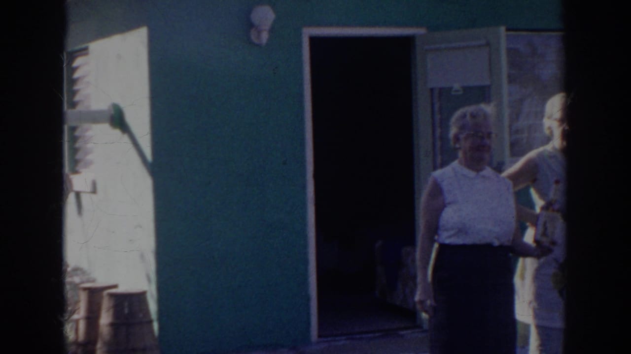 Adults stand near an open doorway on a green building in bright sun.
