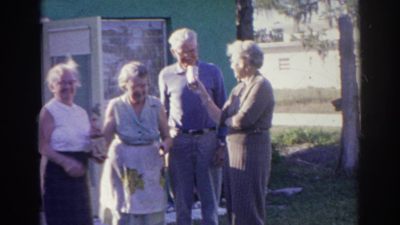 Four older adults stand outside a green house near windows and a tree.