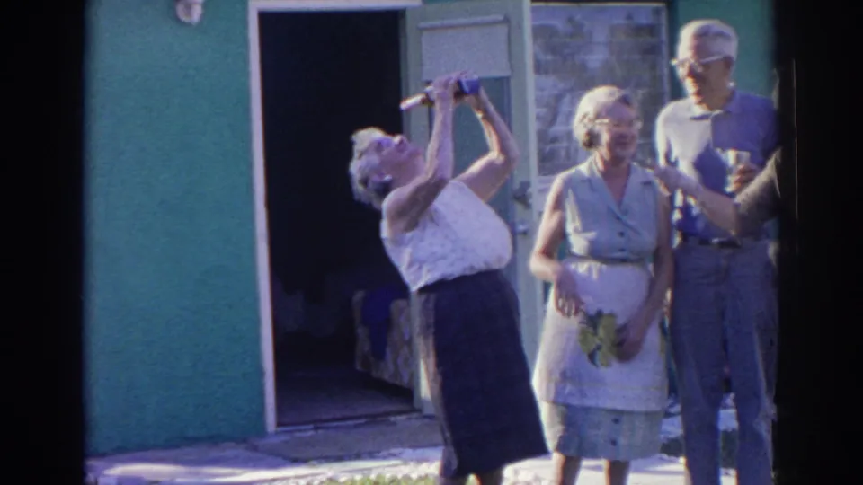 In this 1966 Norfolk home-movie frame, an older woman raises a cup as friends stand nearby outside the house.