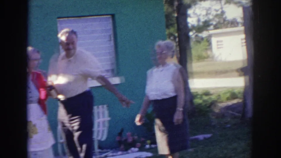 Older friends move through the yard outside the Norfolk house, with a white chair, lawn, and shaded windows visible.