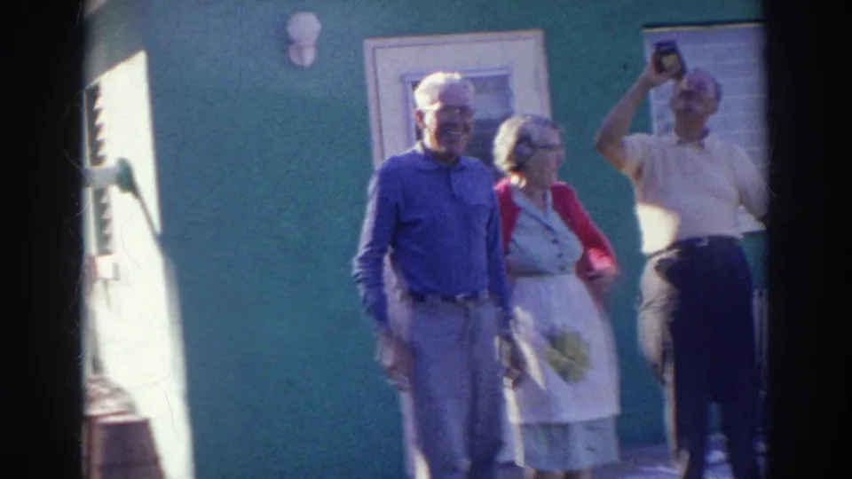 In the Norfolk home-movie scene, three older adults stand in front of the green house while a man lifts a bottle above his head.