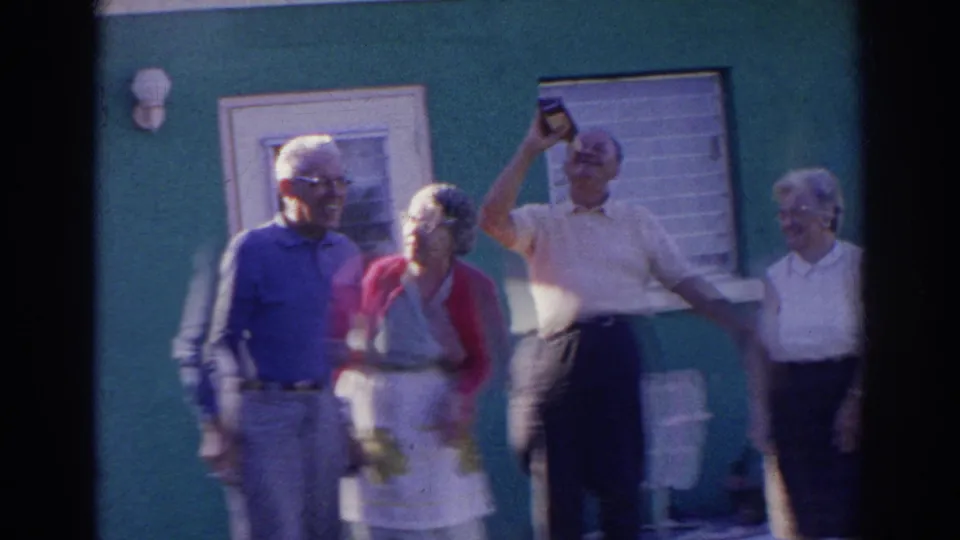 Older adults in 1966 Norfolk stand outside the green house as one man raises a bottle and others look on in a casual moment.
