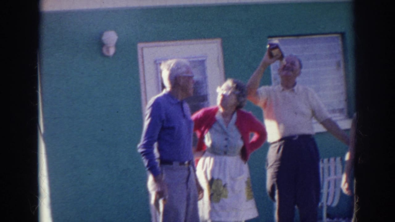 Three older adults watch as a man holds a bottle above his mouth outside a house.