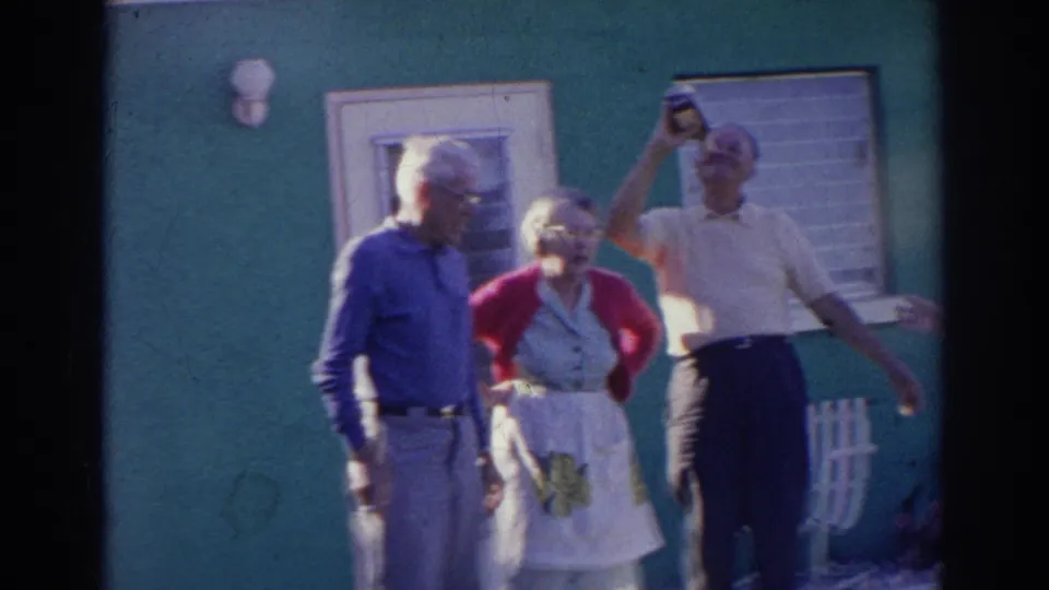 A handheld 8mm frame shows older adults outside the green house as the bottle-drinking gag continues.